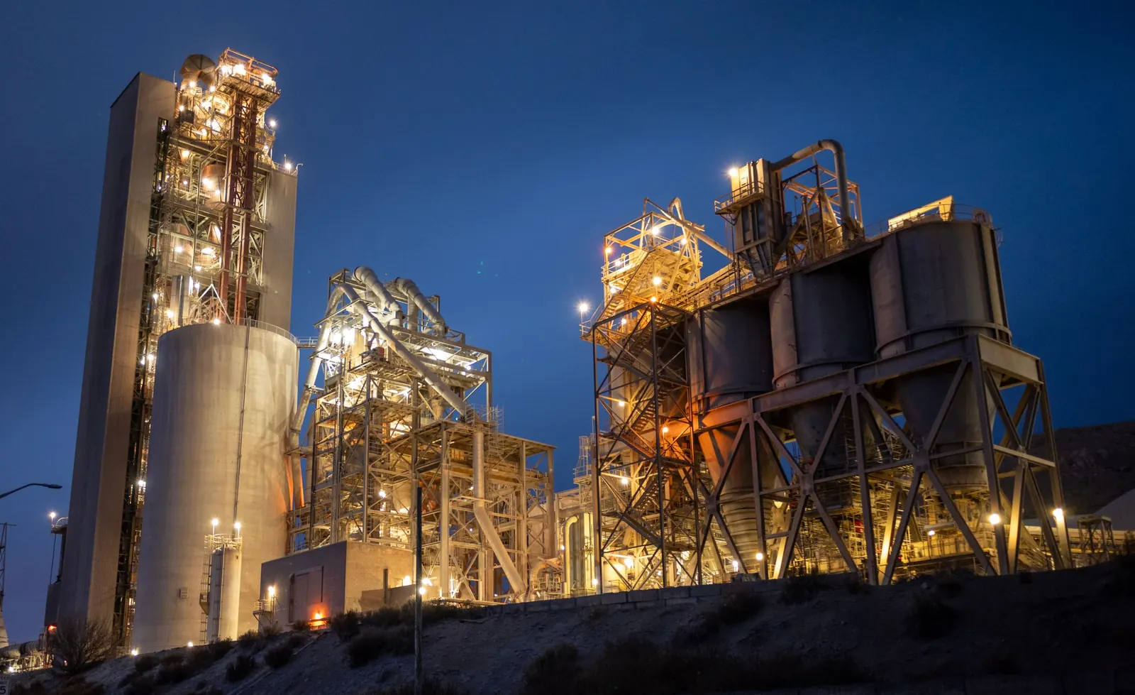 Fábrica de cimento iluminada à noite, com torre de pré-aquecimento, silos e estruturas metálicas