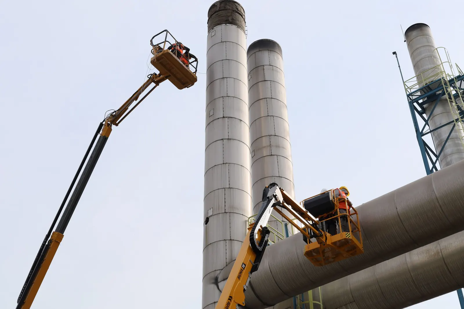 Equipes em plataformas elevatórias fazem manutenção em chaminés e dutos de uma planta industrial